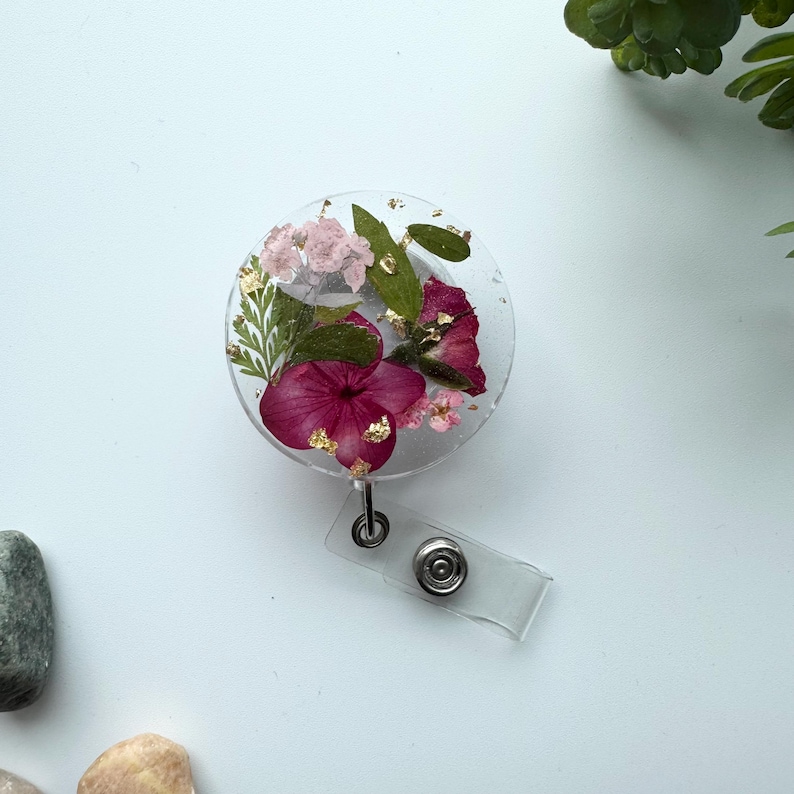 以下が含まれることがあります： ピンクと白の花のデザインと金のアクセントが施された透明な丸いバッジホルダー。バッジホルダーは透明なプラスチック製の伸縮式バッジリールに取り付けられています。
