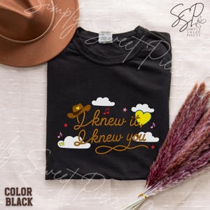 May include: A black t-shirt featuring a graphic with the text "I knew it, I knew you" in a rope-style font. The design includes a brown cowboy hat with a yellow heart, white clouds, a yellow heart, red musical notes, and small red stars.