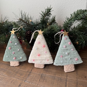 May include: Three triangular ornaments shaped like Christmas trees, two in sage green and one in cream, crafted from textured yarn. Each tree features a small wooden star at the top, a light brown trunk, and scattered pink and white pearl beads.
