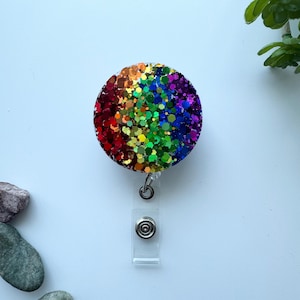 May include: A circular badge reel featuring a vibrant rainbow gradient of chunky hexagonal glitter. The colors transition from deep red and orange on the left to yellow, green, blue, and purple on the right. It includes a clear plastic strap.