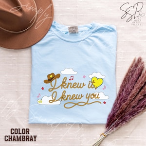 May include: A light blue t-shirt featuring a graphic design with the text "I knew it, I knew you" written in a rope-style font. The design includes a brown cowboy hat, a yellow heart, white clouds, small pink stars, and musical notes.
