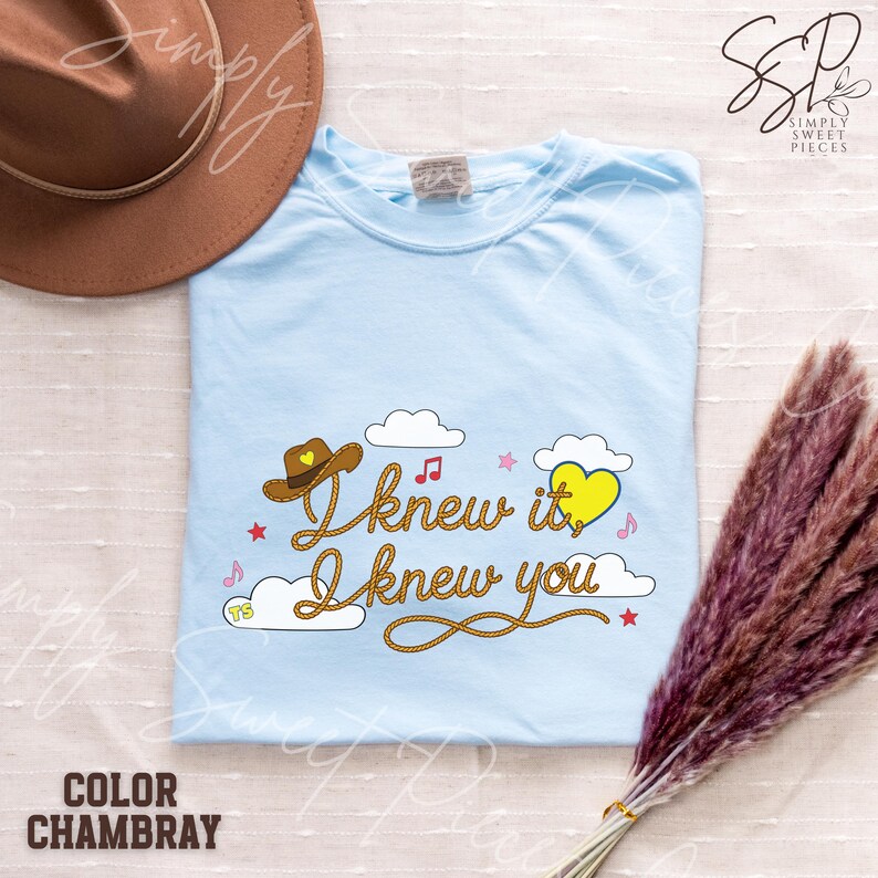 May include: A light blue t-shirt featuring a graphic design with the text "I knew it, I knew you" written in a rope-style font. The design includes a brown cowboy hat, a yellow heart, white clouds, small pink stars, and musical notes.