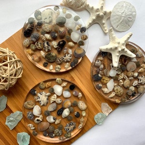 May include: Three round resin coasters with a variety of seashells embedded in the resin. The coasters are on a wooden surface.