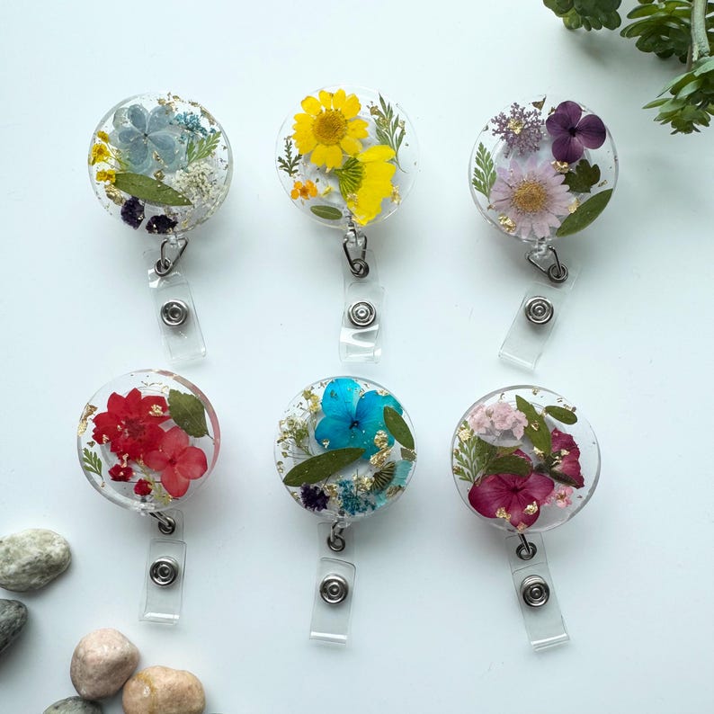 以下が含まれることがあります： 異なる押し花のデザインが施された、透明な樹脂製の丸いバッジホルダー6個。各ホルダーには、シルバーの伸縮式バッジリールが付いています。