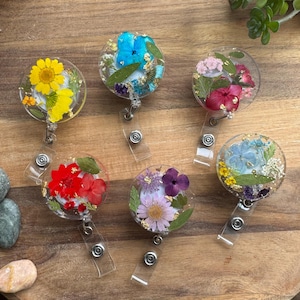 May include: Six round clear resin badge holders with different pressed flower designs. The designs include yellow, red, blue, purple, and white flowers.