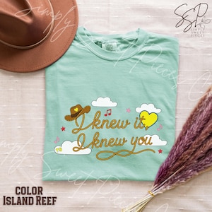 May include: A light green t-shirt featuring the text I knew it, I knew you written in a rope-style font. The design includes a brown cowboy hat with a yellow heart, a yellow heart, white clouds, small pink musical notes, and red stars.