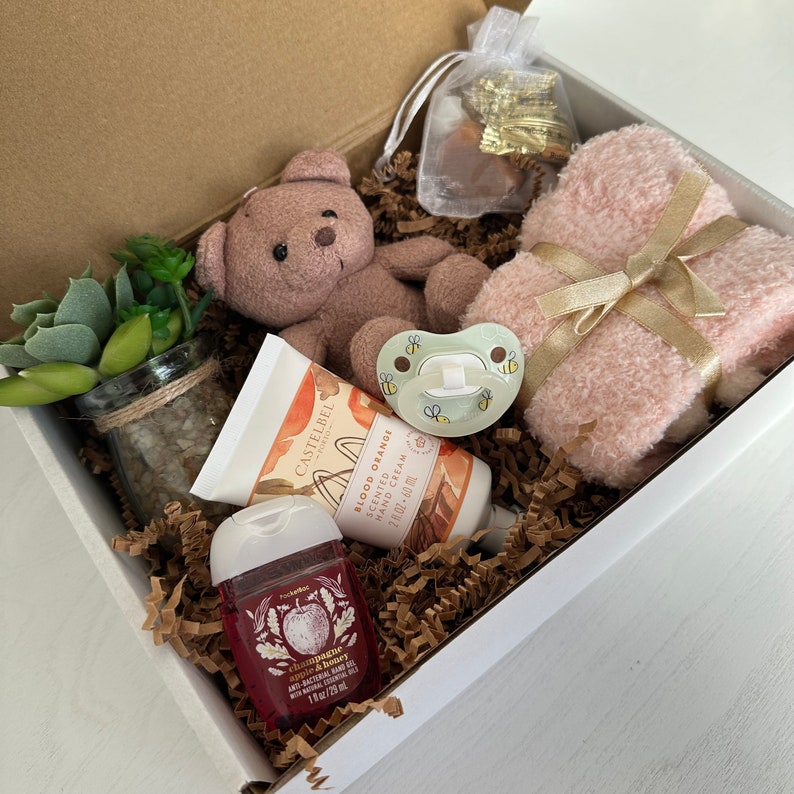 May include: A gift box filled with baby items, including a plush brown teddy bear, a pink towel, a bottle of hand cream, a bottle of hand sanitizer, a small potted succulent, and a pacifier. The box is lined with brown shredded paper.