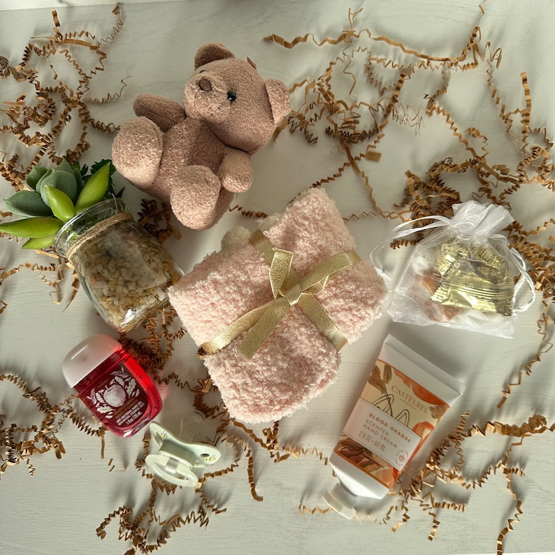May include: A gift basket with a pink fuzzy blanket, a small teddy bear, a bottle of hand cream, a small bottle of hand sanitizer, and a small bag of chocolate candies.