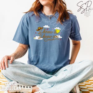 May include: A blue jean colored short-sleeve t-shirt featuring a graphic print with white clouds, a brown cowboy hat, a yellow heart, and the text "I knew it I knew you" in brown cursive lettering, accented by small pink stars and musical notes.