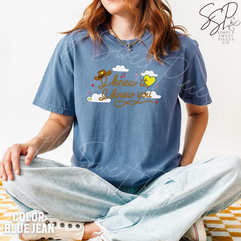 May include: A blue jean colored short-sleeve t-shirt featuring a graphic print with white clouds, a brown cowboy hat, a yellow heart, and the text "I knew it I knew you" in brown cursive lettering, accented by small pink stars and musical notes.