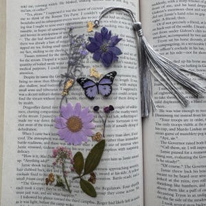 May include: A handmade bookmark featuring pressed purple flowers, a butterfly, and a silver tassel. The bookmark is placed on a book page with text, creating a delicate and artistic accessory for book lovers.