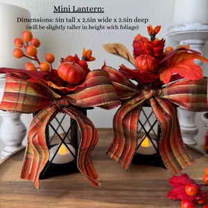 May include: Two black mini lanterns, each adorned with a plaid ribbon bow and autumn foliage. The lanterns are 5 inches tall, 2.5 inches wide, and 2.5 inches deep. The text on the image reads "Mini Lantern: Dimensions: 5in tall x 2.5in wide x 2.5in deep (will be slightly taller in height with foliage)".