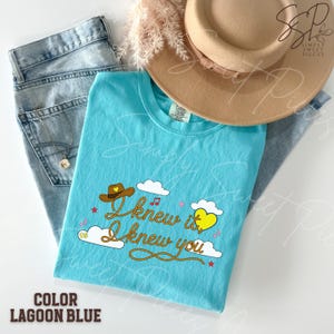 May include: A lagoon blue t-shirt featuring a graphic with a brown cowboy hat, a yellow heart, white clouds, and red musical notes. The text reads I knew it, I knew you in a rope-style font. The shirt is paired with blue denim jeans and a tan hat.