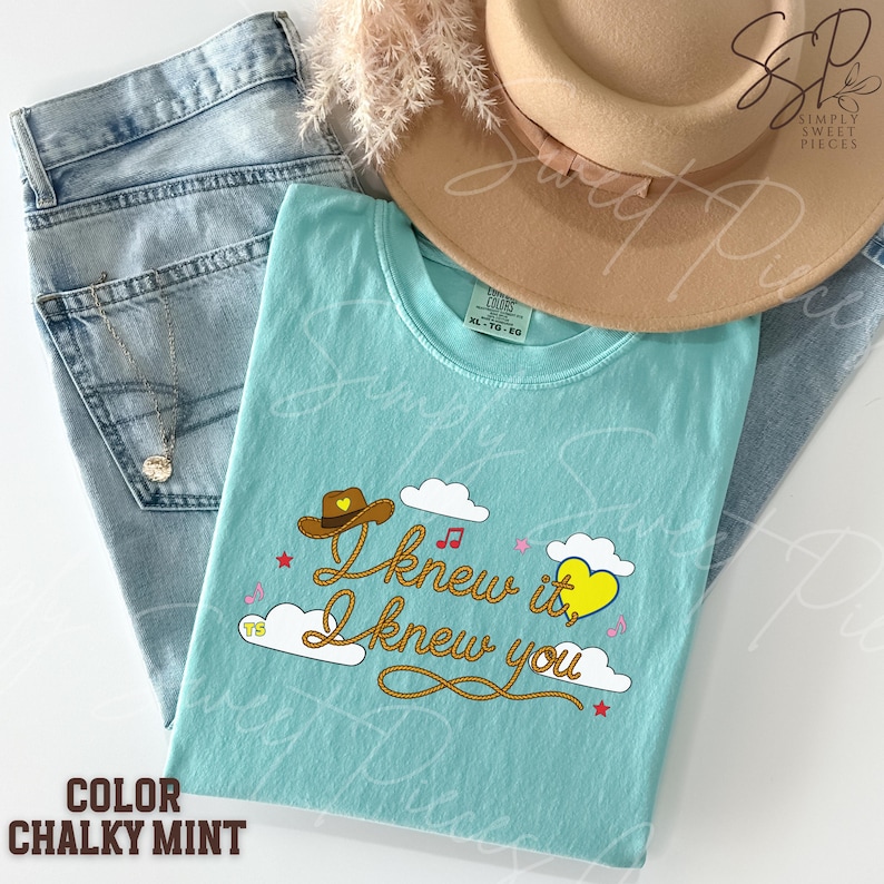May include: A light mint green t-shirt featuring a graphic with the text I knew it, I knew you in a rope-style font. The design includes a brown cowboy hat with a yellow heart, white clouds, small red stars, and pink musical notes.