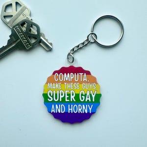 May include: A keychain with a rainbow-colored, scalloped-edge pendant. The pendant has the text "COMPUTA, MAKE THESE GUYS SUPER GAY AND HORNY" in white. A silver key ring and chain are attached to the pendant. Two keys are visible in the background.