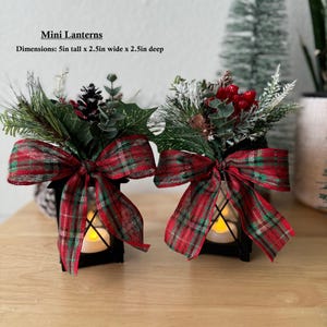 May include: Two black metal lanterns with red and green plaid bows and greenery. The lanterns are 5 inches tall, 2.5 inches wide, and 2.5 inches deep.  Mini Lanterns.