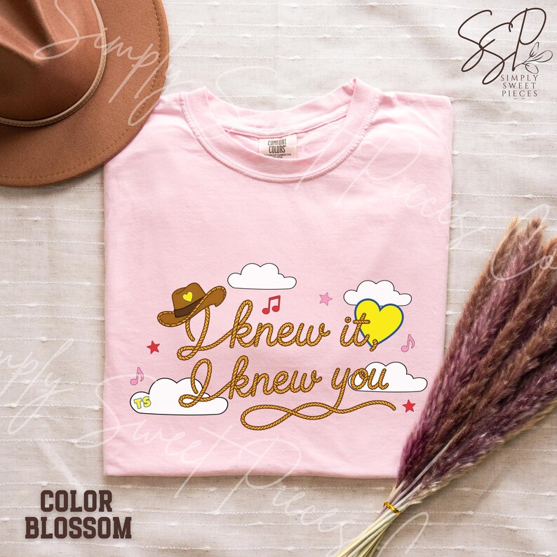 May include: A light pink t-shirt featuring a graphic with the text I knew it, I knew you written in a rope-style font. The design includes a brown cowboy hat with a yellow heart, white clouds, small pink stars, musical notes, and a yellow heart.
