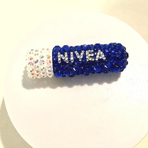 May include: A blue and white Nivea lipstick tube covered in sparkling crystals. The blue section features the brand name in silver letters. The white end is also covered in crystals. The lipstick is displayed on a white surface.