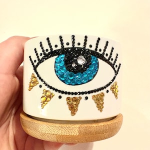 Puede incluir: Una maceta de cerámica blanca con un diseño de ojo turco azul y negro. La maceta tiene un borde de purpurina dorada y una base de bambú.
