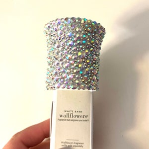 Può includere: Un diffusore di fragranze per pareti bianco con una parte superiore trasparente e iridescente ricoperta di cristalli. Il diffusore ha il testo "White Barn wallflowers* Fragrance that welcomes you home*" sul davanti. Il testo "Wallflowers fragrance refills sold separately" è in basso.