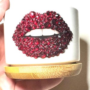 May include: A white ceramic planter with a red rhinestone lip design. The planter has a bamboo base.