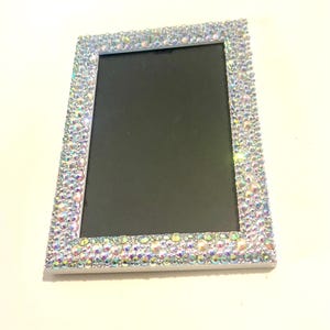 May include: Rectangular photo frame with a black interior and a white border. The frame is heavily embellished with iridescent, sparkling rhinestones, creating a glamorous and eye-catching design. The rhinestones are closely set, covering the entire frame.