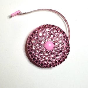 May include: Pink measuring tape with a round, pink, rhinestone-covered case. The case has a small, pink button in the center.