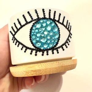 May include: White ceramic planter with a blue evil eye design made of rhinestones. The planter has a bamboo base.