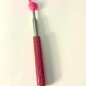 May include: A pink selfie stick with a rhinestone-covered handle and a pink hand-shaped phone holder. The extendable silver pole is topped with a bright pink hand gesture, perfect for taking photos.