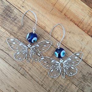 May include: Silver butterfly earrings with a dark blue bead featuring a white and light blue eye design. The earrings have a delicate, openwork design. The earrings are displayed on a wooden surface.