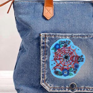 Peut inclure: Un sac fourre-tout en jean avec un écusson décoratif. L'écusson est de forme irrégulière bleu clair avec un motif de toile d'araignée rouge et noir, entouré de fleurs bleues et de verdure. Le sac a une poignée en cuir marron.