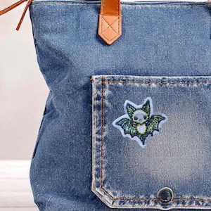 May include: A blue denim tote bag with a brown leather handle and a bat patch on the front pocket. The bat patch features a white skull and green wings with a white border. The bag is made of denim.