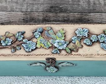 Puede incluir: Una caja de madera rectangular pintada a mano, con base turquesa y tapa crema. La tapa presenta un diseño en relieve de un pájaro, flores y mariposas en tonos azules, verdes y marrones. La caja tiene un cierre metálico.