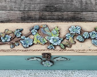 Puede incluir: Una caja de madera decorativa con tapa con bisagras. La tapa está pintada con un diseño floral con flores azules, hojas verdes y un pájaro. La caja está pintada en tonos azules y crema.