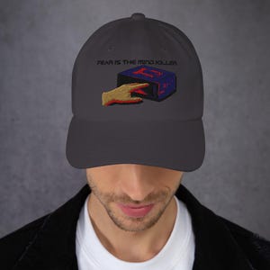 May include: A dark gray baseball cap with an embroidered design featuring a hand reaching towards a blue and red box with the text "FEAR IS THE MIND KILLER".