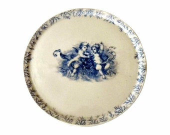 May include: A white ceramic plate with a blue and white floral border and a central design of two cherubs in blue.