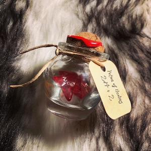 Könnte beinhalten: Eine kleine Glasflasche mit einem Korkverschluss und einem roten Wachssiegel. Die Flasche enthält einen roten 20-seitigen Würfel und ein Etikett mit der Aufschrift "Trank der Heilung 2d4 + 2".