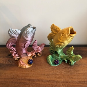 以下が含まれることがあります： 魚の形をした3Dプリントのサイコロタワーが2つ。左側のタワーはピンクとシルバーの魚で、中に青と金のサイコロが入っています。右側のタワーは金の魚で、中に緑と金のサイコロが入っています。