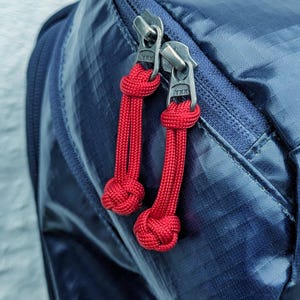 Könnte beinhalten: Nahaufnahme eines marineblauen Rucksacks mit zwei silbernen Reißverschlüssen. Jeder Reißverschluss hat eine leuchtend rote Kordel mit einem Knoten. Die Reißverschlüsse sind mit "YKK" gekennzeichnet.