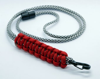 Könnte beinhalten: Ein rotes Paracord-Lanyard mit einem schwarz-weiß gemusterten Band und einem schwarzen Metallclip.