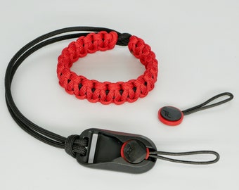 May include: A red and black paracord camera wrist strap with a black plastic quick release buckle. The strap has a small red and black button on a black cord.