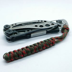 Puede incluir: Una multiherramienta plateada con mango negro y un cordón de paracord verde y rojo. La multiherramienta tiene el texto "LEATHERMAN" y "Skeleteool" impreso en ella.