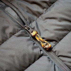 May include: Close-up of a black quilted jacket with a zipper. The zipper pull is a multi-colored cord in orange, black, and yellow. The jacket has a diagonal quilted pattern.