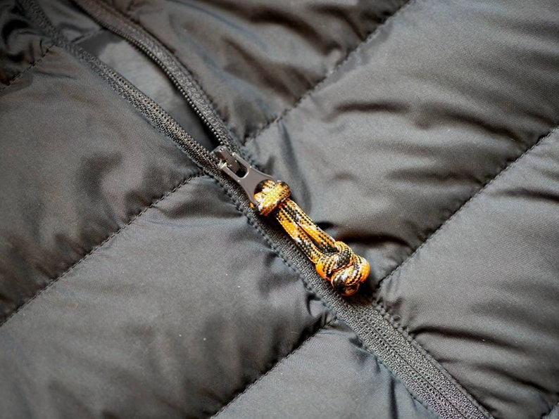 May include: Close-up of a black quilted jacket with a zipper. The zipper pull is a multi-colored cord in orange, black, and yellow. The jacket has a diagonal quilted pattern.