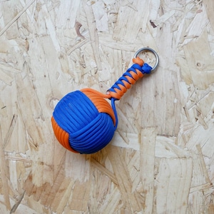 Puede incluir: Un llavero de paracord azul y naranja con un anillo de metal. El llavero está atado en un nudo para formar una bola.