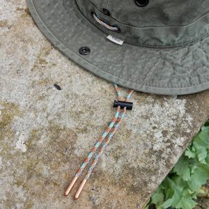 May include: An olive green hat with a wide brim, featuring a small white tag with the text Tilley. The hat includes a multi-colored patterned cord with a black plastic sliding toggle and two metallic copper-colored aglets at the ends of the cord.