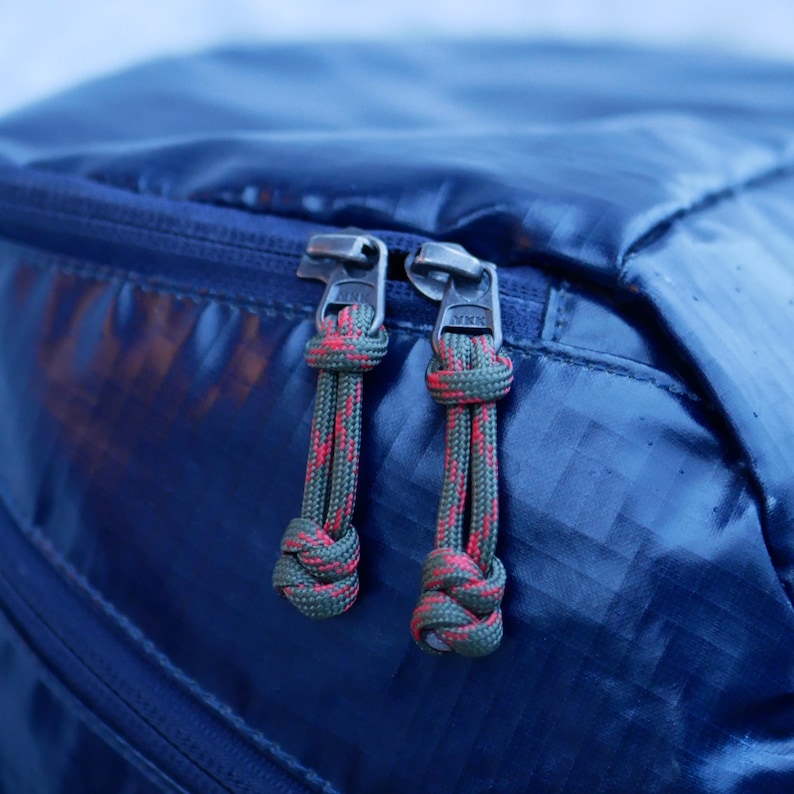 May include: Close-up of a navy blue backpack with two zippers. Each zipper has a pull made of green and red rope. The zippers are silver and have the letters "YKK" printed on them.