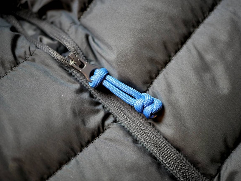 May include: Close-up of a black quilted jacket zipper with a blue cord pull. The zipper is black metal, and the jacket fabric has a subtle sheen. The blue cord is knotted for easy gripping.