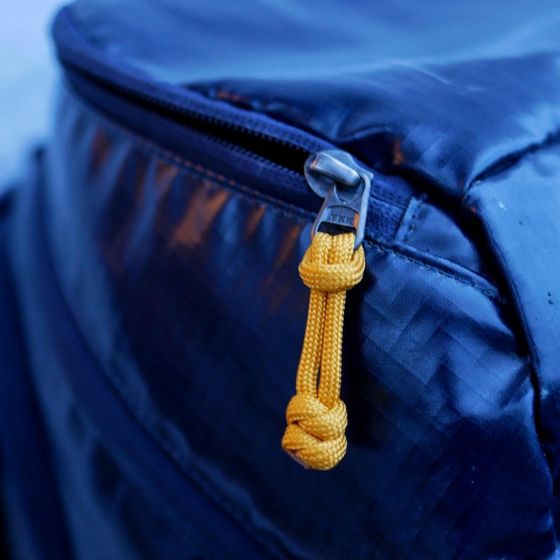 May include: Close-up of a blue backpack with a silver zipper and a yellow cord pull. The zipper is partially open, revealing the interior. The cord pull is knotted and provides easy access to the zipper.