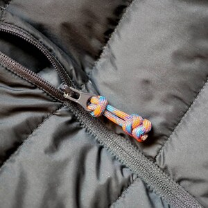 May include: Close-up of a black quilted jacket zipper with a colorful, braided pull. The zipper is closed, and the pull features a mix of blue, orange, and yellow cords. The jacket's fabric has a diamond pattern.